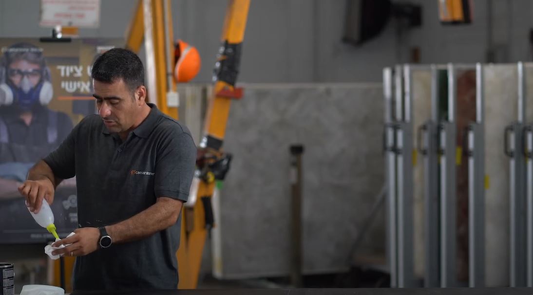 עובד במפעל קוורץ מכין חומר בעזרת בקבוק סחיטה, כשהוא לובש חולצת עבודה של Caesarstone וברקע ציוד תעשייתי ולוחות קוורץ מסודרים