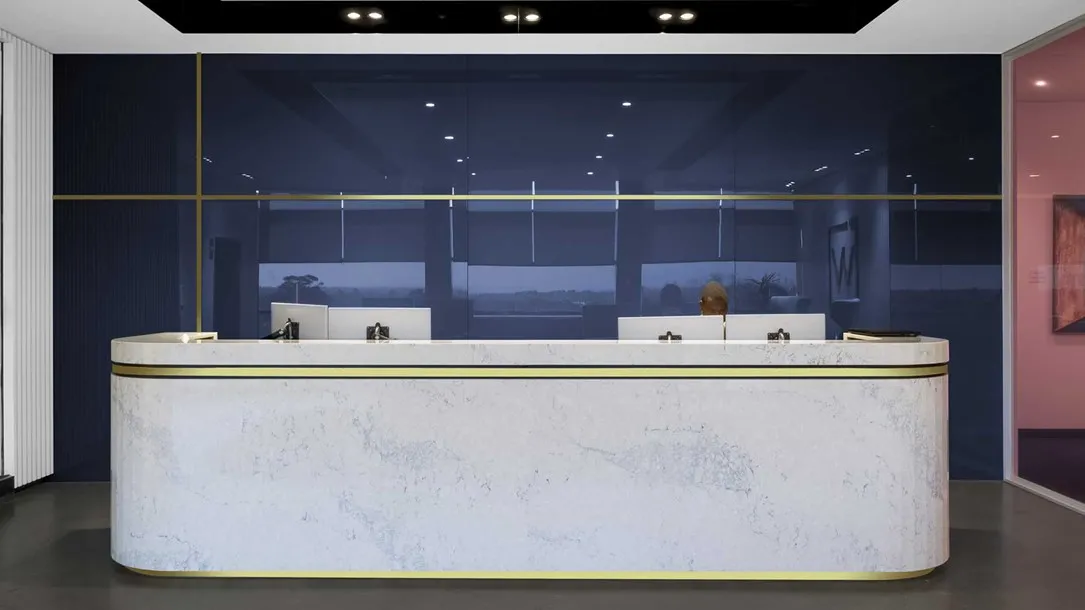 דלפק קבלה מסחרי עשוי אבן קיסר בגוון בהיר עם כיפופי-חום (Thermoforming), בחזית חלל מודרני עם קיר זכוכית כהה ותאורה שקועה.